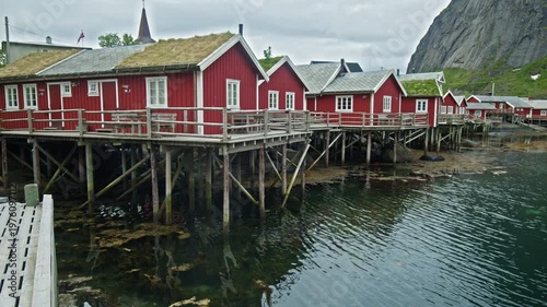 Row of red rorbuer cabins on stilts above water in Lofoten, Norway. Classic fishing village with reflections and mountain backdrop.