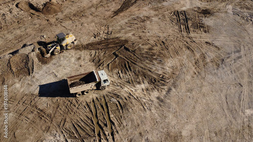 A high-angle aerial shot captures heavy machinery operating on a dirt construction site. A yellow front-end loader is shown filling the bed of a large dump truck with soil and debris.