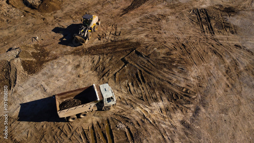A high-angle aerial shot captures heavy machinery operating on a dirt construction site. A yellow front-end loader is shown filling the bed of a large dump truck with soil and debris.