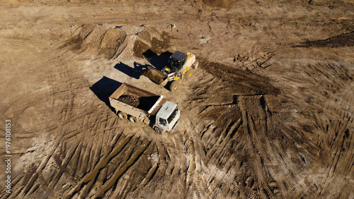 A high-angle aerial shot captures heavy machinery operating on a dirt construction site. A yellow front-end loader is shown filling the bed of a large dump truck with soil and debris.