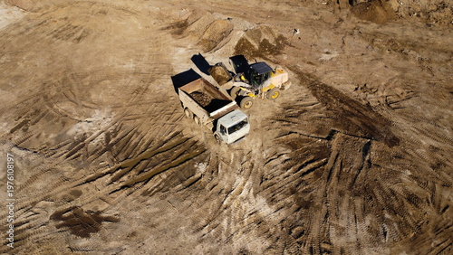 A high-angle aerial shot captures heavy machinery operating on a dirt construction site. A yellow front-end loader is shown filling the bed of a large dump truck with soil and debris.
