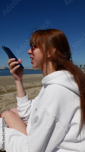Young woman dictating text with AI voice chat on smartphone by the sea.