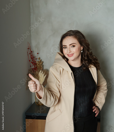 Confident woman with long wavy brown hair wearing a stylish beige hooded coat shows thumbs up gesture and smiles while posing in a modern minimalist office with gray walls and decorative elements