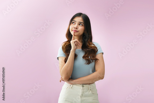 Indonesian woman touching chin while looking upward with thoughtful and curious expression, searching for ideas or answers, isolated on pink background