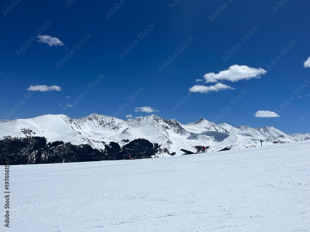 Fototapeta premium Wide view of a snow-covered mountain range and ski slope under a deep blue sky with scattered white clouds on a clear winter day.