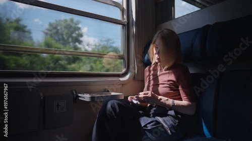 Relaxed mature woman with blonde hair is sitting by the window in a train compartment. She is putting in her wireless earbuds and enjoying the sunny scenery passing by