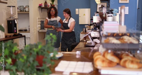 Two diverse female employees working at a local coffee shop, using a digital tablet to manage orders and inventory, showcasing teamwork and modern small business operations