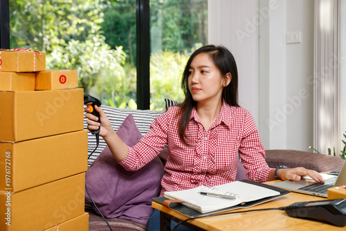 Young Asian SME owner scanning barcode on parcel box for delivery. E-commerce woman checking stock inventory at home office. Logistics and online selling process concept.