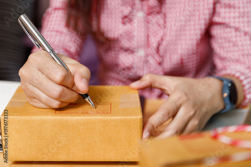 Close up of online seller hand writing address on cardboard box with pen. Small business owner preparing parcel for shipping. E-commerce delivery concept.