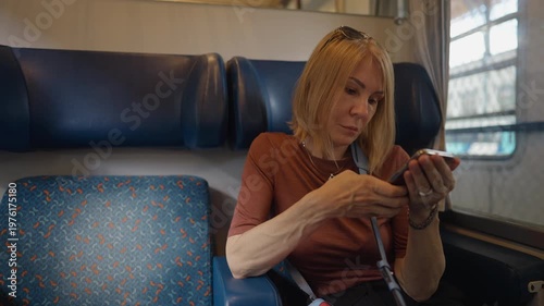Mature blonde woman sitting by the window in a train compartment, focused on her smartphone screen while traveling. Commuting and technology concept