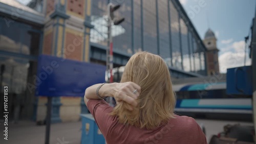 Rear view of a woman with blonde hair walking towards Nyugati railway station in Budapest as a train passes by. She adjusts her hair, windblown
