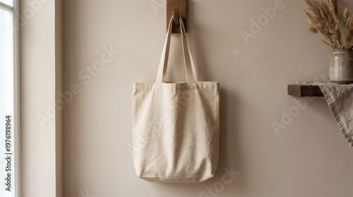 Photograph of a beige canvas tote bag hanging on a wooden wall hook, next to a window with natural light. A wooden shelf with dried wheat stalks and a woven basket is also visible in the background.