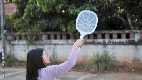 Close-up of a woman using an electric mosquito swatter outdoors in a residential area. Suitable for pest control, hygiene, and daily life themes.