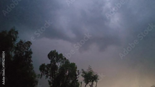 Dramatic lightening & thunder clouds loom over silhouetted trees as the sky transitions from dark to lighter shades of gray and blue