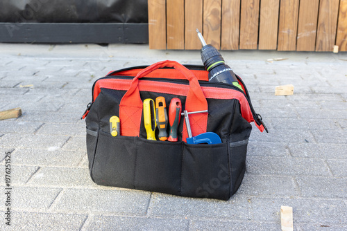 Tool bag with tools on a construction site resting on the pavement near a wooden wall