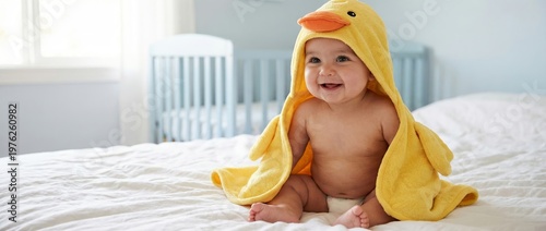 A baby sits on a bed wearing a yellow duck towel hood