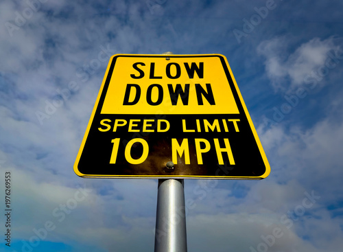Картината върху платно A yellow sign in a parking lot stating Slow Down Speed Limit 10 MPH against a bl