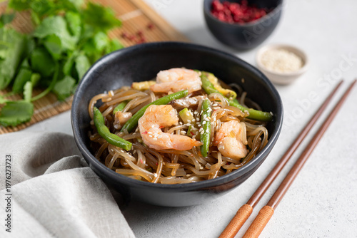 Asian glass noodles with shrimps and green beans served in bowl on grey background, garnished with sesame seeds. Wooden chopsticks on side
