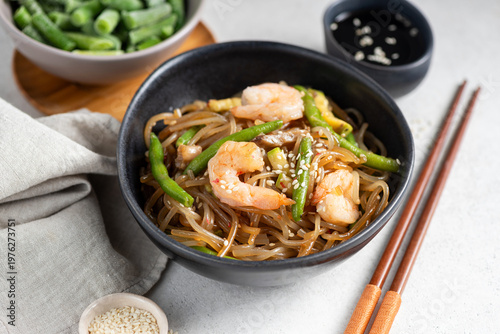 Asian glass noodles with shrimps and green beans served in bowl on grey background, garnished with sesame seeds. Wooden chopsticks on side