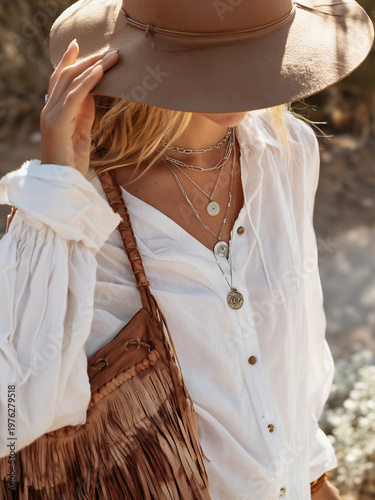 A woman wearing a brown hat and layered necklaces while holding a fringed bag.
