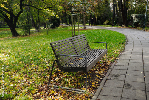 Wooden park bench near paved walkway surrounded by green grass and trees in peaceful outdoor park setting.