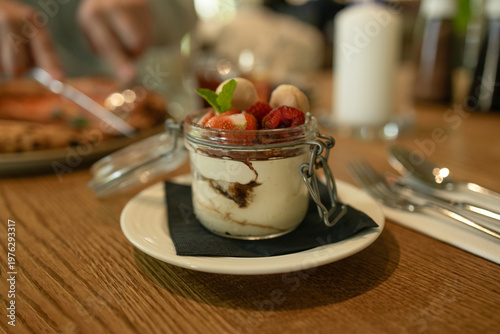 Delicious layered dessert with fresh strawberries and cream served in glass jar on wooden table in cozy cafe setting.