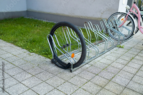 A white bicycle is secured to a metal bike rack on a tiled sidewalk. Colorful artwork adorns the nearby building, creating a vibrant urban atmosphere in the bright daylight. High quality photo