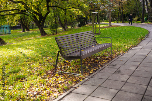 Wooden park bench near paved walkway surrounded by green grass and trees in peaceful outdoor park setting.