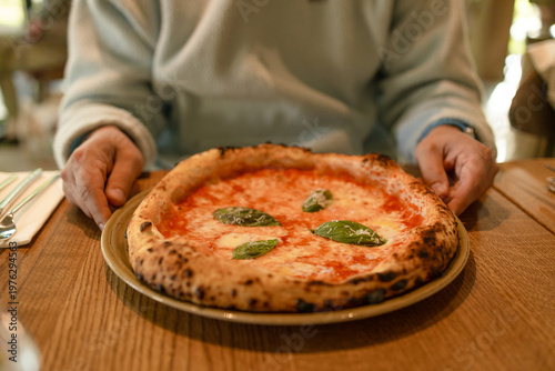 Hands serving freshly baked margherita pizza with tomato sauce and basil on wooden table in cozy restaurant setting.