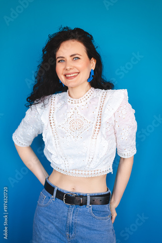 Smiling young woman with curly dark hair posing with hands on hips against bright blue studio background happy female in white lace top looking at camera with joyful expression commercial portrait