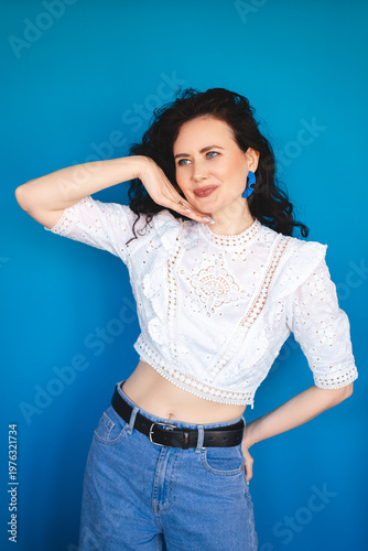 Smiling young woman with curly dark hair posing with hand near face against bright blue studio background happy female in white lace top looking away with joyful expression commercial portrait shot