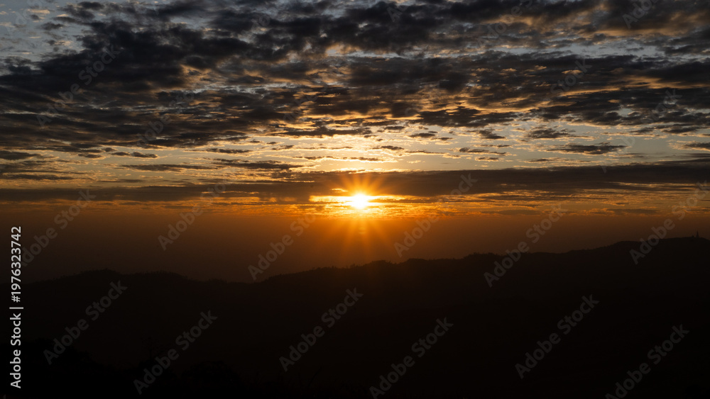 Obraz premium Majestic Golden Sunburst Breaking Through Dramatic Clouds over Mountain Silhouettes at Sunrise