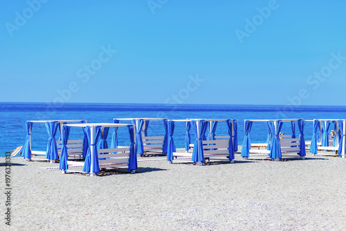 White beach cabanas with blue curtains on a sandy shore under a clear blue sky, summer vacation concept. Copy space, sea resort landscape.