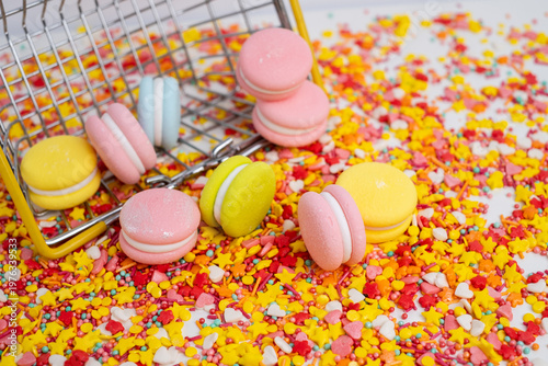 Miniature supermarket basket with colorful macarons spilled on bright candy sprinkles background. Sweet shopping and food consumption concept. 