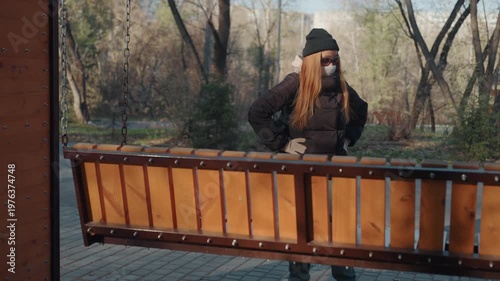 White female prowler at park bench, masked figure in winter coat and beanie, hands on hips in tense stance near wooden swing suspended by chains, empty autumn park with trees and paved path, morning