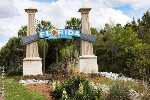 Florida Welcomes You Sign Stands at Roadside Under Cloudy Sky With Palm Trees and Plants in the Background