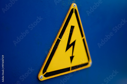 Yellow triangular high voltage warning sign with lightning symbol mounted on blue metal surface in close up.