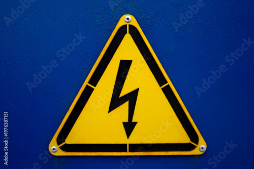 Yellow triangular high voltage warning sign with lightning symbol mounted on blue metal surface in close up.