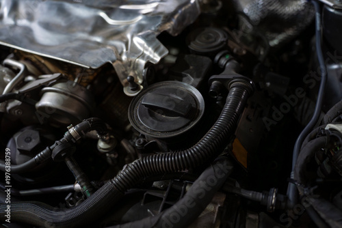 Close view of a car engine bay with oil filler cap, hoses, wiring, and metal components under the hood in workshop lighting.