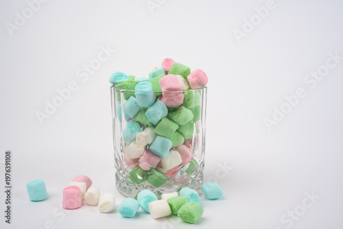 Assorted pastel marshmallows fill a clear textured glass, spilling onto a bright white surface. This arrangement creates a sweet, playful scene in a well-lit indoor environment.