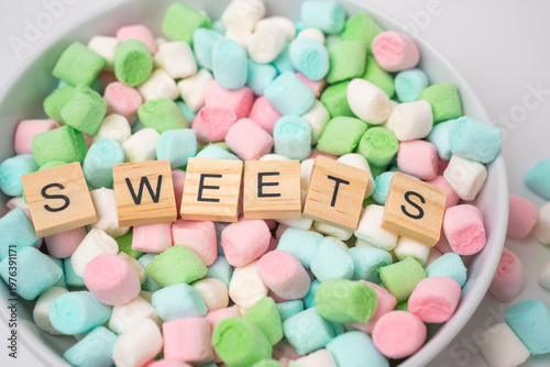 A white bowl holds a vibrant mix of pink, green, blue, and white mini marshmallows. Wooden letter tiles arranged on top spell out the word sweets, creating a playful and colorful dessert display