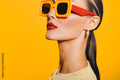 Fotografi Stylish woman with slicked back hair wears large orange sunglasses and red lipstick against bright yellow background