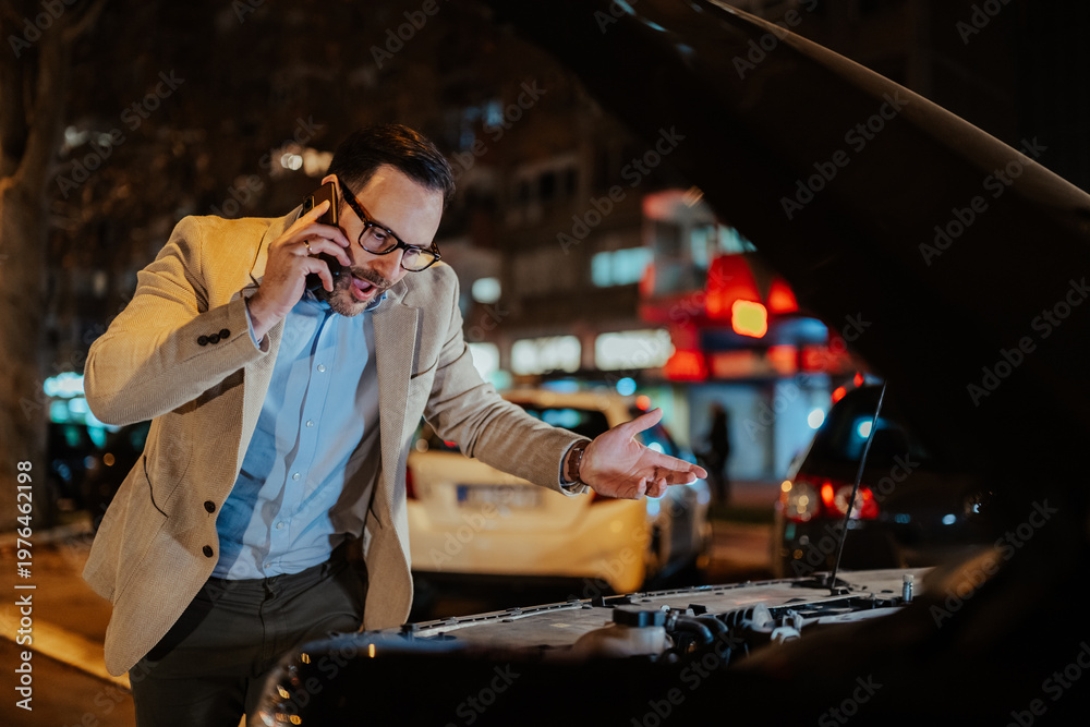 Obraz premium Man talking on smartphone, standing by his broken car on a city street at night, engine hood open, needing help