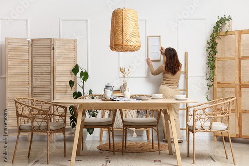 Young woman hanging blank frame on light wall in dining room, back view
