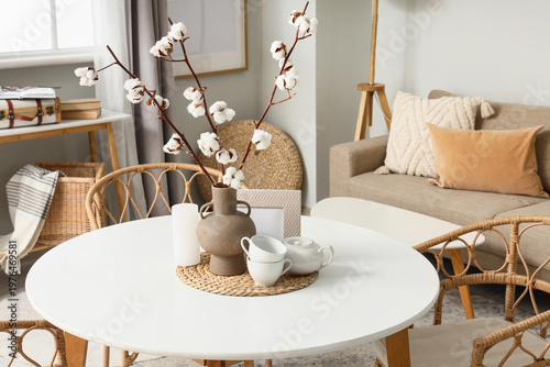 Vase with cotton flowers, burning candle and tea set on dining table in room