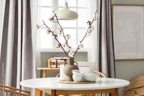 Vase with cotton flowers, burning candle and tea set on dining table in room