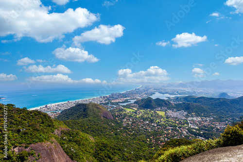Breathtaking panoramic view of Barra da Tijuca, Rio de Janeiro, from a mountain peak. Features lush green mountains, blue ocean, coastal city skyline, and lagoons under a sunny sky with clouds.