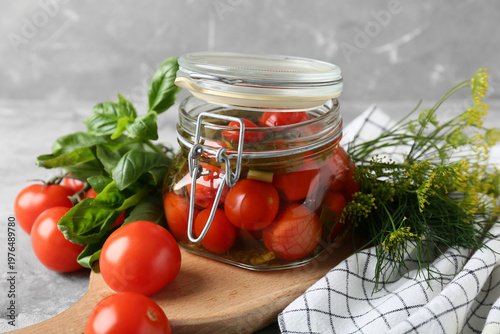 Jar of tasty canned tomatoes with dill and basil leaves on grey background
