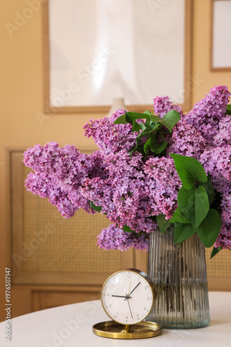 Vase with lilac flowers and alarm clock on table in living room, closeup