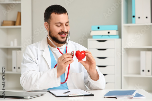 Young happy male doctor with stethoscope and red heart sitting at table in cl...